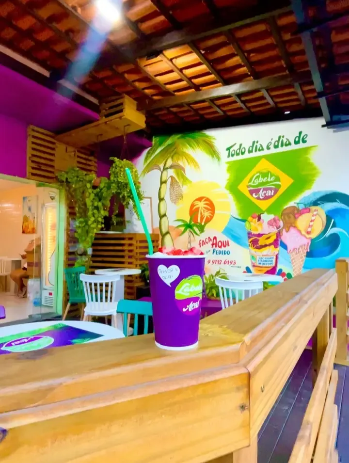 O salão da Labele: mural de coqueiros com Todo dia é dia de e o logo, plantas, mesas brancas e um copo roxo no balcão